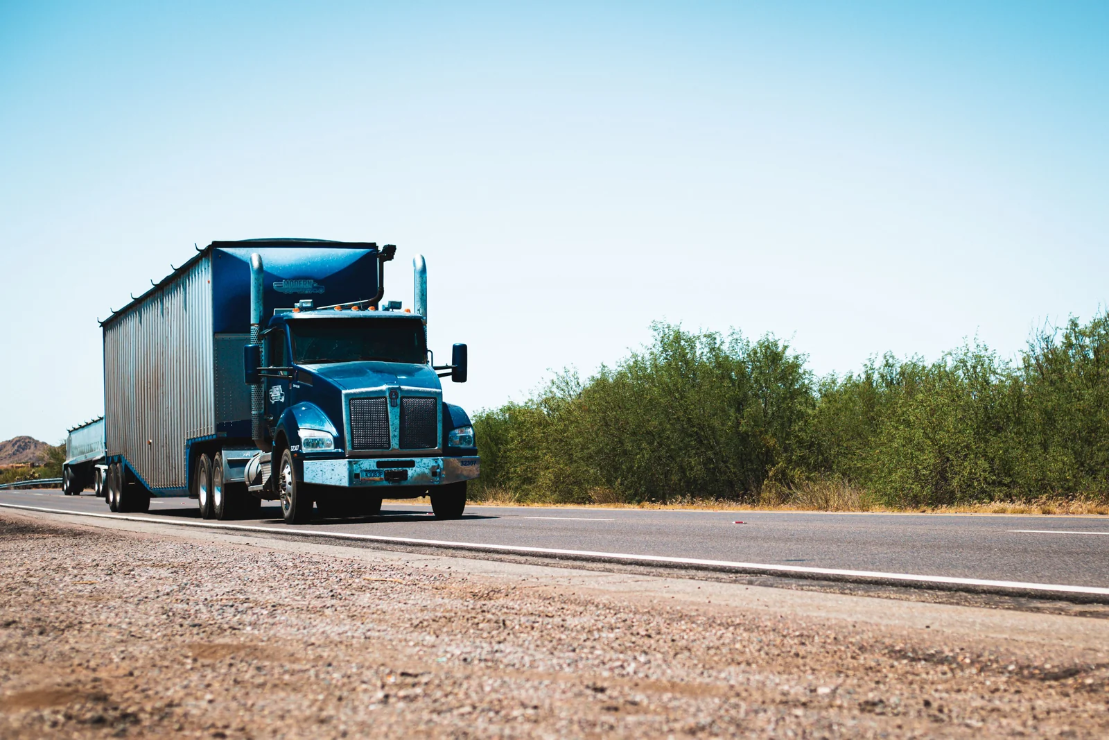 A blue freight truck travelling on a highway under a clear sky.